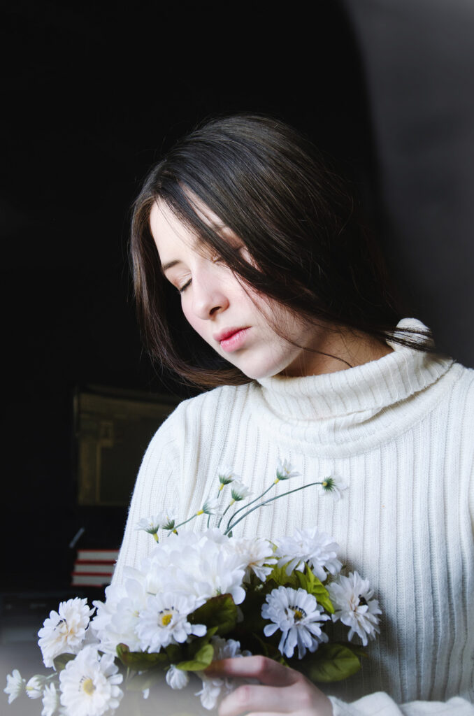 Gabriela Inzunza a serene individual with long dark hair is captured in a contemplative pose holding a bouquet of white flowers against a dark backdrop.